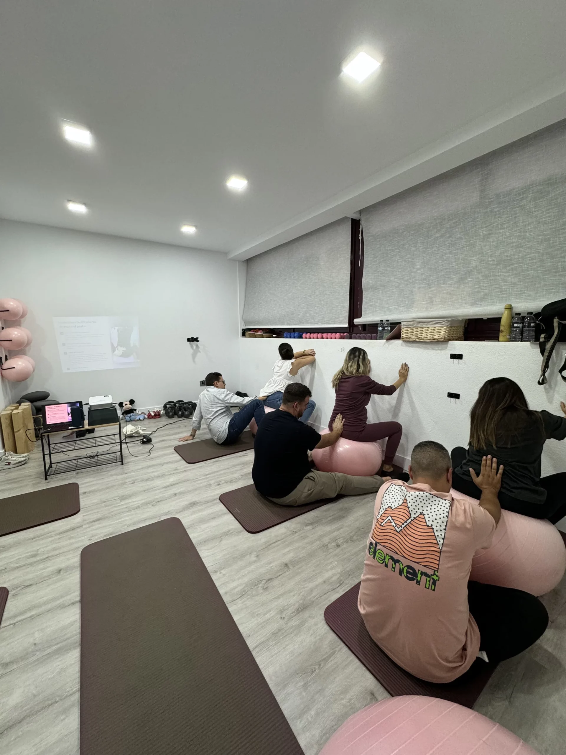 Un grupo de seis personas, incluyendo parejas, asisten a un taller de preparación para el parto en una sala bien iluminada. Las personas están sentadas en colchonetas de yoga y utilizando grandes pelotas de ejercicio (fitballs) de color rosa para practicar posturas y ejercicios de movimiento. Una pequeña proyección en la pared blanca muestra texto e imágenes. Se ven algunos equipos como un ordenador portátil y material de yoga a la izquierda.