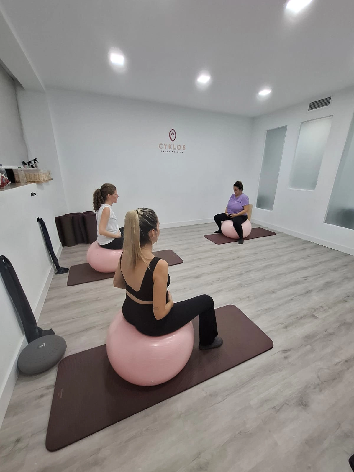 Tres mujeres, embarazadas, están sentadas sobre grandes balones de ejercicio de color rosa pálido (fitballs o balones suizos) en una sala de ejercicios moderna. Están sentadas en colchonetas de color burdeos. Dos de las mujeres se ven de espaldas o de perfil, y una tercera mujer se ve de frente, sentada más atrás en la sala. En la pared blanca central, hay un logotipo de color burdeos que dice "CYKLOS SALUD PÉLVICA". La sala está bien iluminada y tiene un suelo de madera clara.