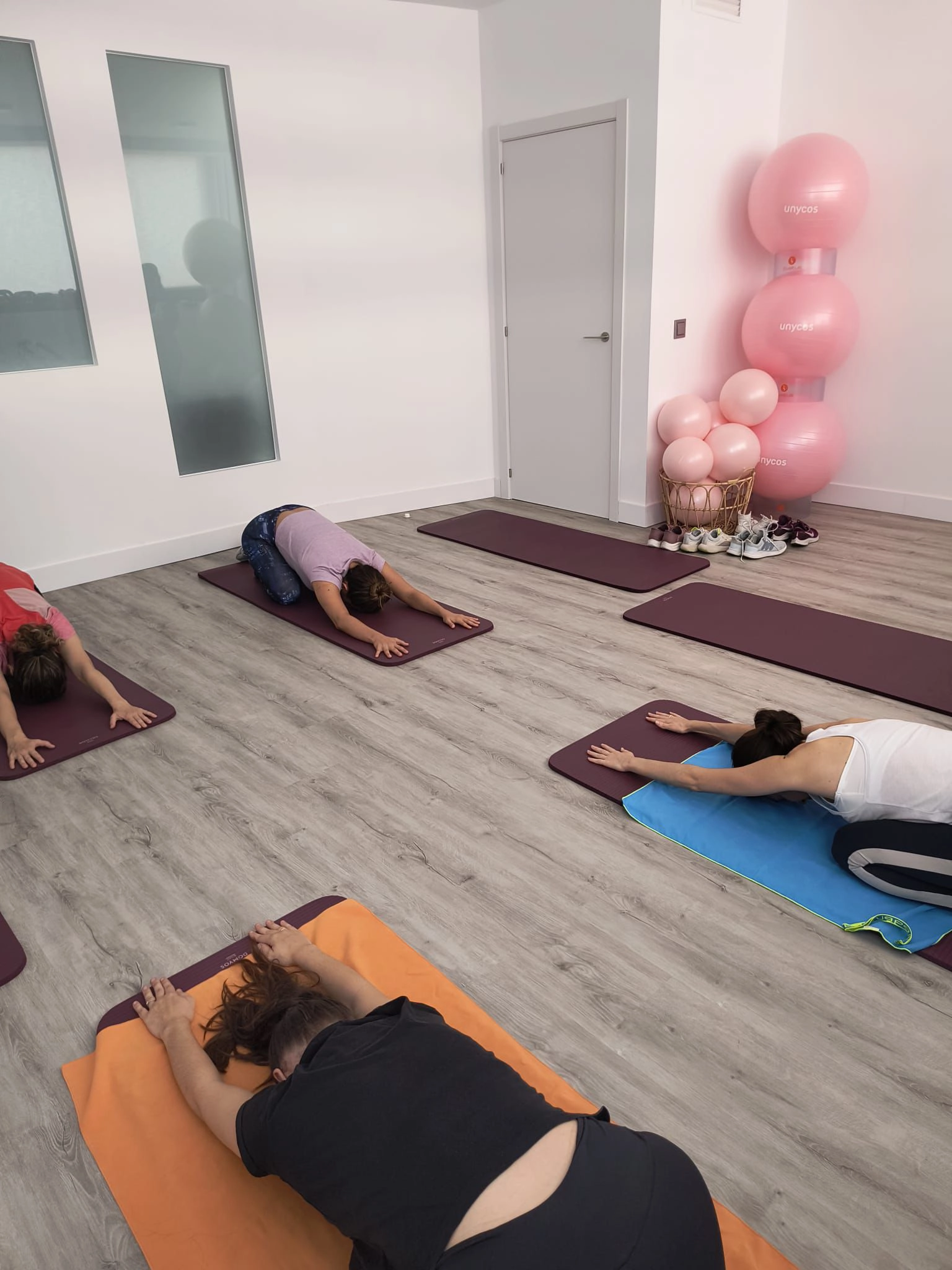 Cuatro personas se encuentran en una luminosa sala de ejercicios o estudio de yoga, ejecutando la postura del niño (Balasana) sobre colchonetas individuales de color burdeos. El suelo es de tablones de madera clara. En la esquina superior derecha, se apilan varias pelotas de ejercicio grandes de color rosa con el logo 'unycos', y debajo hay una cesta con zapatillas deportivas. La sala tiene paredes blancas y un espejo de pared con acabado esmerilado parcial.