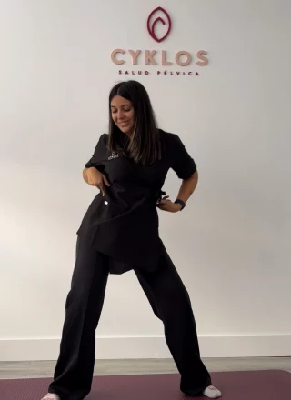 Una mujer joven, con cabello oscuro y vestida con un uniforme sanitario negro de corte cruzado y pantalones a juego, posa de manera enérgica y divertida sobre una colchoneta de ejercicios color burdeos. Está inclinada con las piernas separadas y las manos en la cintura o apuntando a su abdomen, mirando hacia abajo con una sonrisa. Detrás de ella, en una pared blanca, se ve el logotipo de color burdeos y cobre que dice "CYKLOS SALUD PÉLVICA".