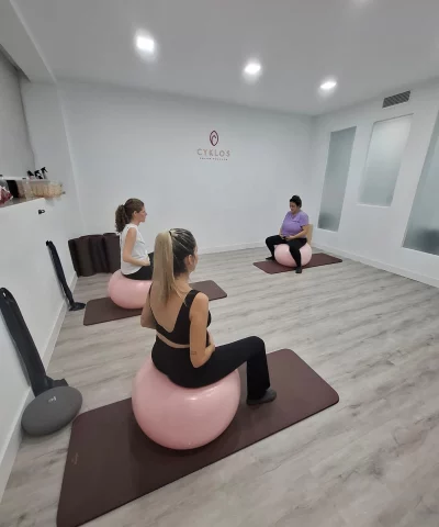 Tres mujeres, embarazadas, están sentadas sobre grandes balones de ejercicio de color rosa pálido (fitballs o balones suizos) en una sala de ejercicios moderna. Están sentadas en colchonetas de color burdeos. Dos de las mujeres se ven de espaldas o de perfil, y una tercera mujer se ve de frente, sentada más atrás en la sala. En la pared blanca central, hay un logotipo de color burdeos que dice "CYKLOS SALUD PÉLVICA". La sala está bien iluminada y tiene un suelo de madera clara.